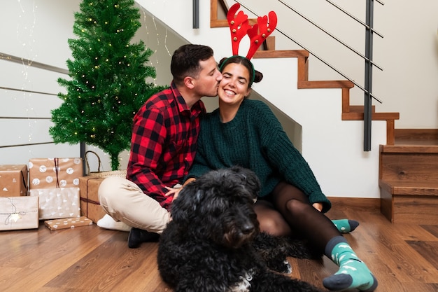 Jovem kisse a namorada na bochecha para o Natal, usando chifres de rena, cachorro preto, feriado