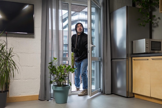 Jovem ladrão em roupas escuras entra com cuidado e silêncio pela janela da varanda no prédio de apartamentos Segura acessórios de sucata