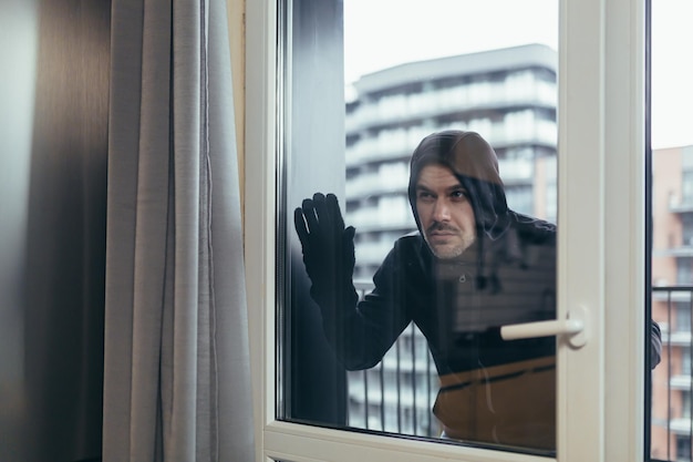 Jovem ladrão masculino em roupas escuras olha pela janela do apartamento Ele quer entrar em habitação