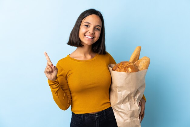 Jovem Latina comprando alguns pães isolados no azul mostrando e levantando um dedo em sinal dos melhores