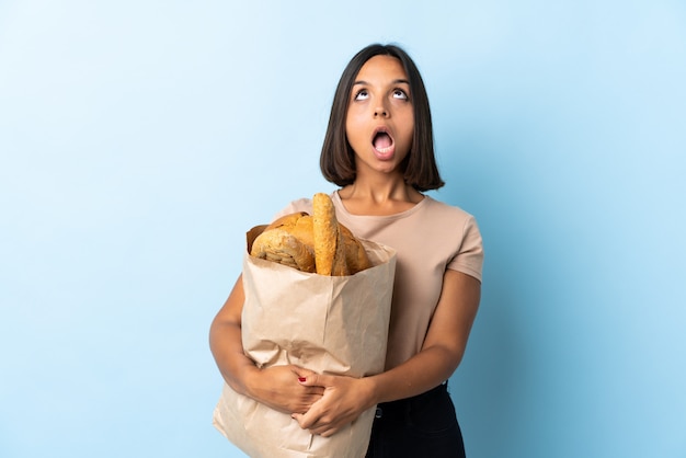 Jovem Latina comprando alguns pães isolados no azul, olhando para cima e com expressão de surpresa