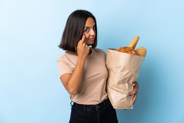 Jovem latina comprando pães isolados em azul mostrando algo