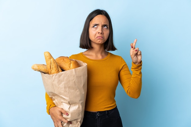 Jovem latina comprando pães isolados no azul com os dedos se cruzando e desejando o melhor