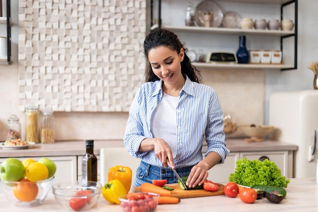 Jovem latina cozinhando salada deliciosa e saudável em pé na cozinha moderna cortando pepino em