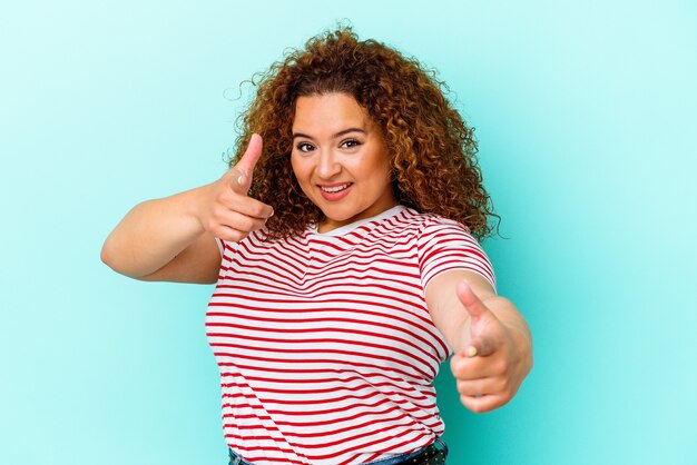 Jovem latina curvilínea isolada na parede azul, apontando para a frente com os dedos.