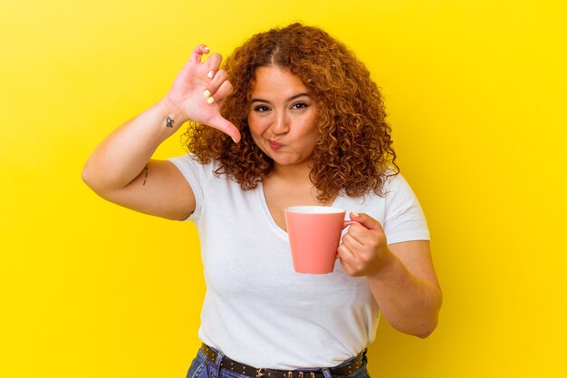 Jovem latina curvilínea segurando uma xícara isolada em um fundo amarelo, mostrando um gesto de antipatia, polegares para baixo. Conceito de desacordo.