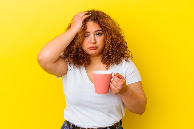 Jovem latina curvilínea segurando uma xícara isolada em um fundo amarelo, sendo chocada, ela se lembrou de uma reunião importante.