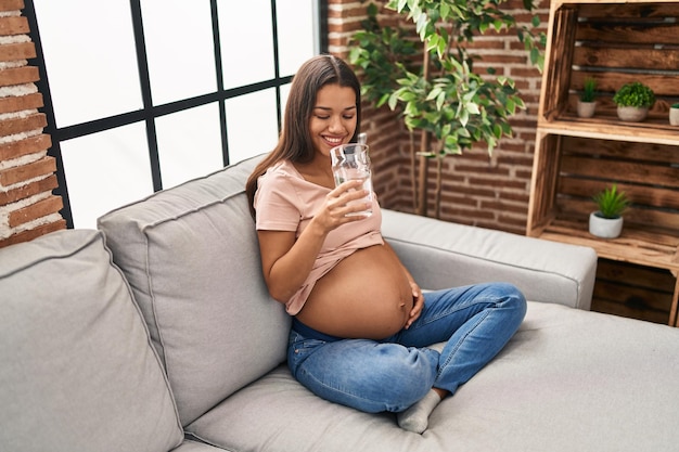 Jovem latina grávida bebendo água sentada no sofá em casa
