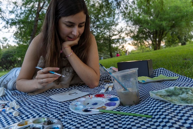 Jovem latina pintando com aquarela em um piquenique no parque cercado por grama e árvores