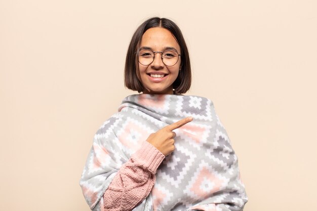 Foto jovem latina sorrindo alegremente, sentindo-se feliz e apontando para o lado e para cima, mostrando o objeto no espaço da cópia