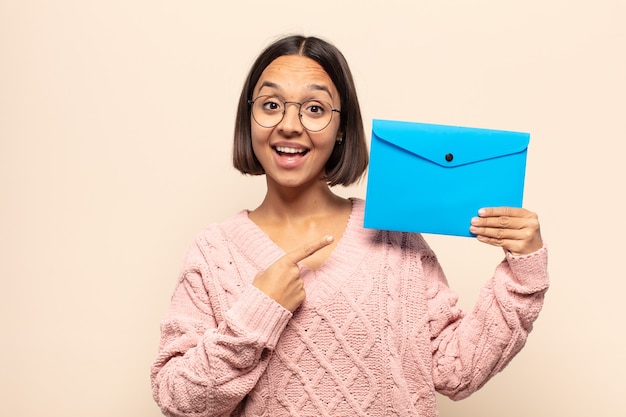 Jovem latina sorrindo alegremente, sentindo-se feliz e apontando para o lado e para cima, mostrando o objeto no espaço da cópia