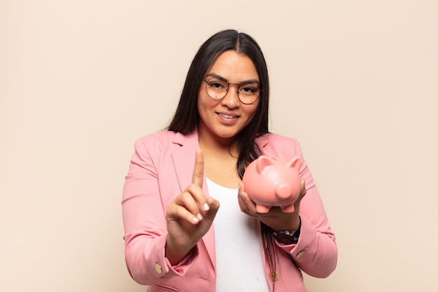 Jovem latina sorrindo com orgulho e confiança fazendo a pose número um triunfantemente, sentindo-se uma líder