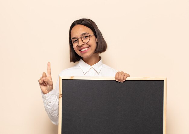 Jovem latina sorrindo com orgulho e confiança fazendo a pose número um triunfantemente, sentindo-se uma líder