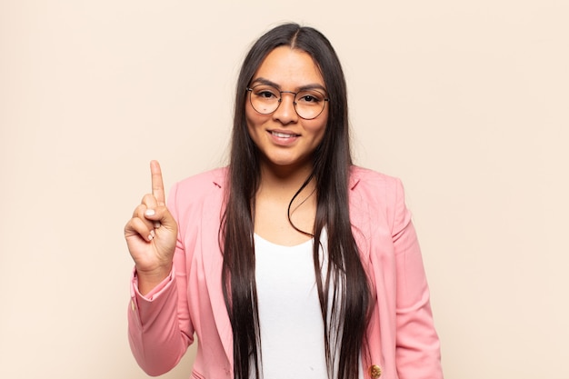 Jovem latina sorrindo e parecendo amigável, mostrando o número um ou primeiro com a mão para a frente, em contagem regressiva