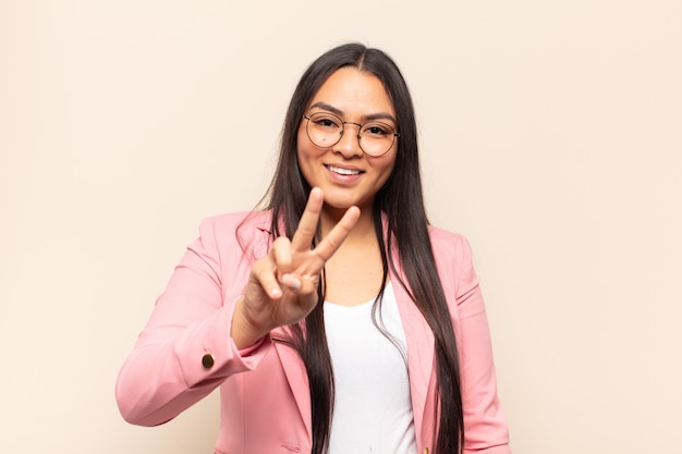 Jovem latina sorrindo e parecendo feliz, despreocupada e positiva, gesticulando vitória ou paz com uma mão