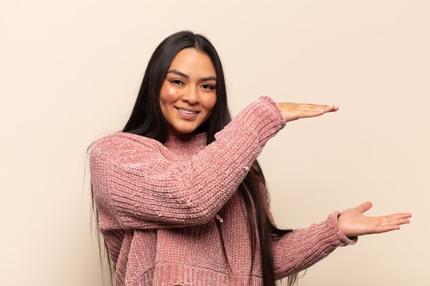 Jovem latina sorrindo, sentindo-se feliz, positiva e satisfeita, segurando ou mostrando um objeto ou conceito no espaço da cópia