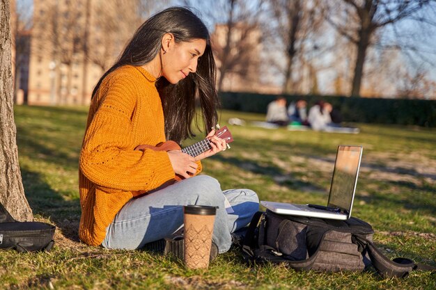 Jovem latina tocando ukulele em um parque ela faz uma pausa na cidade praticando em um parque com seu instrumento musical olhando as letras no laptop