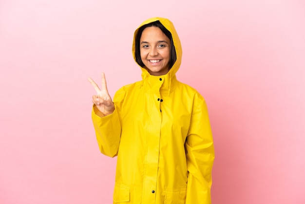 Jovem latina vestindo um casaco à prova de chuva sobre um fundo isolado, sorrindo e mostrando o sinal da vitória