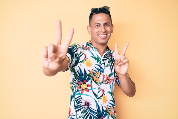 Jovem latino bonito vestindo roupas de verão sorrindo olhando para a câmera mostrando os dedos fazendo o sinal de vitória número dois