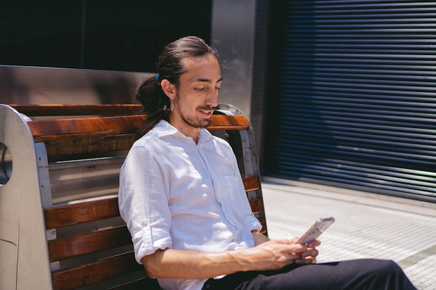 Jovem latino com cabelo comprido e barba vestindo uma camisa fazendo uma videochamada de um banco da cidade Copiar espaço