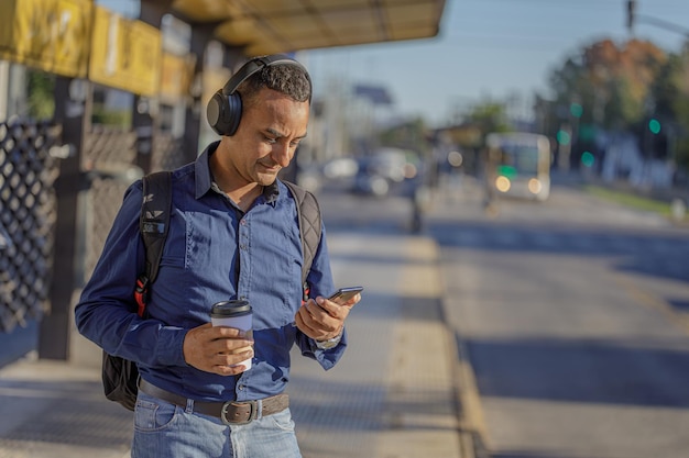Jovem latino com fones de ouvido olhando para seu celular no ponto de ônibus