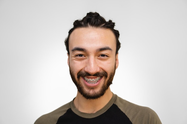 Jovem latino e hispânico com aparelho e barba sorrindo enquanto olha para a câmera com cabelo trançado em um fundo branco