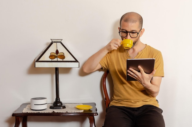 Jovem latino em roupas casuais e óculos lendo em um tablet enquanto bebe café sentado em uma cadeira em casa Copie o espaço