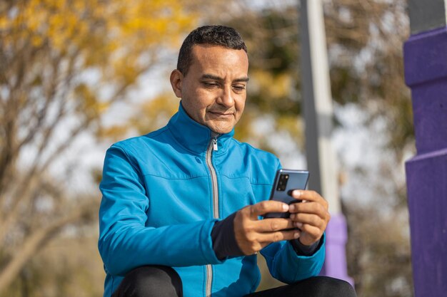 Jovem latino em roupas esportivas olha para seu celular sentado em algumas escadas em um parque público