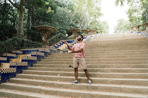 Jovem latino fazendo malabarismo nas ruas ensolaradas de Barcelona fazendo um show com algumas bolas brancas enquanto sorria Vestido com calças marrons e uma camiseta listrada pastel