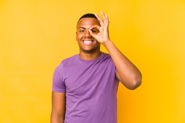 Jovem latino isolado em amarelo animado, mantendo o gesto ok no olho.