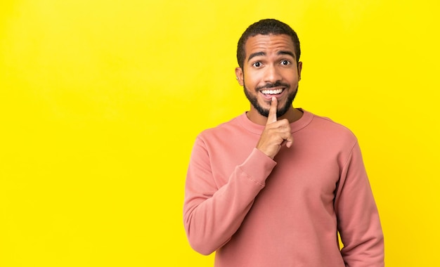 Jovem latino isolado em fundo amarelo, mostrando um sinal de gesto de silêncio, colocando o dedo na boca