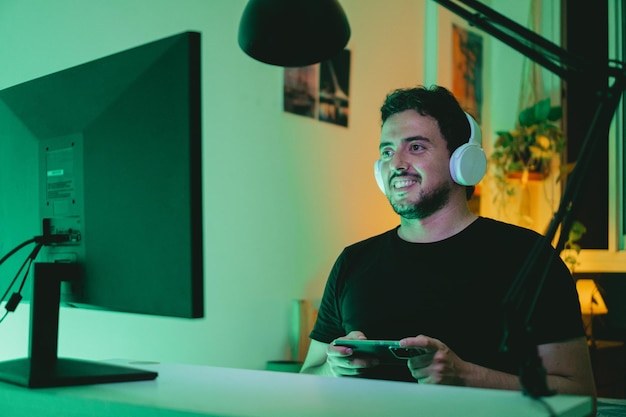 Foto jovem latino jogando videogame em seu computador de mesa enquanto usa fones de ouvido e usa seu celular como joystick