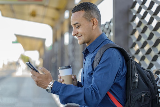 Jovem latino olhando para seu celular com um copo de papel com café na mão