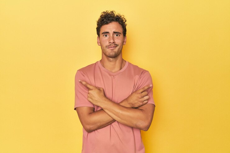 Foto jovem latino posando em fundo amarelo aponta para o lado está tentando escolher entre duas opções