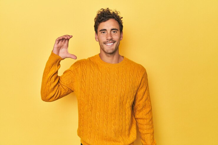Foto jovem latino posando em fundo amarelo segurando algo pequeno com os dedos indicadores sorrindo