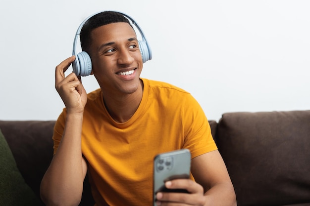 Jovem latino positivo vestindo roupas casuais ouvindo música em fones de ouvido segurando telefone celular