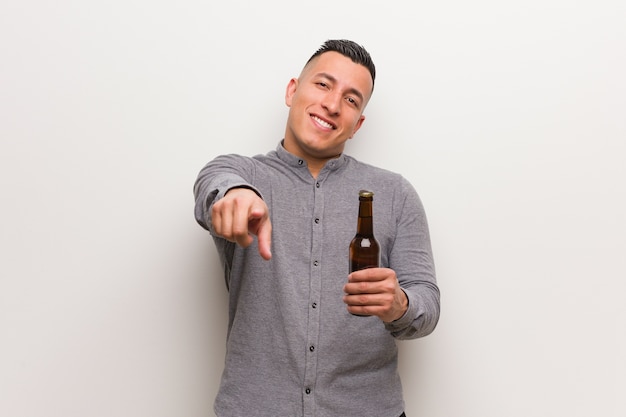 Foto jovem latino segurando uma cerveja, alegre e sorridente, apontando para a frente