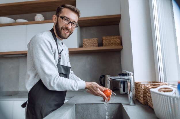 Jovem lavando tomates sob a torneira na cozinha