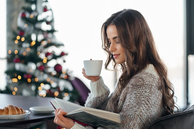 Jovem lendo livro perto da árvore de natal em casa