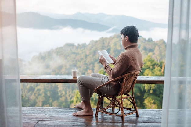 Jovem lendo livro perto da janela e olhando para a montanha em casa de família rural ao nascer do sol da manhã. SoloTravel, viagem, viagem e conceito relaxante