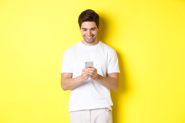 Jovem lendo mensagem de texto no smartphone, olhando para a tela do celular e sorrindo, usando uma camiseta branca contra a parede amarela