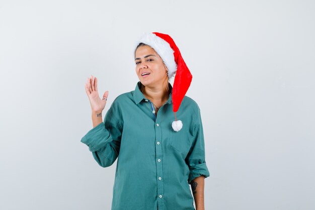 Jovem, levantando a mão com chapéu de Natal, camisa e olhando alegre. vista frontal.