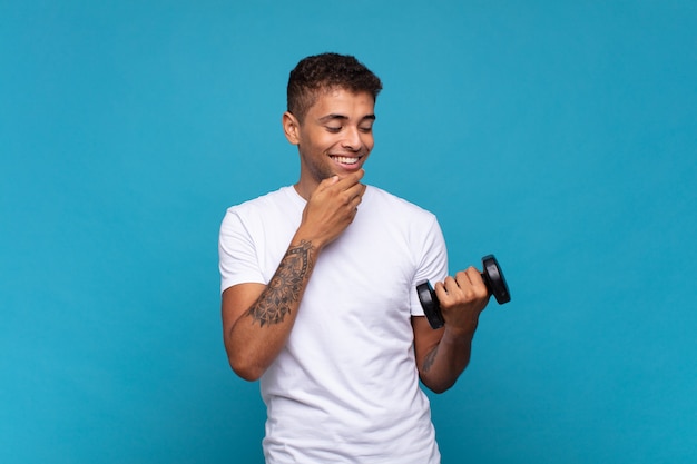 Jovem levantando um haltere sorrindo com uma expressão feliz e confiante com a mão no queixo, pensando e olhando para o lado