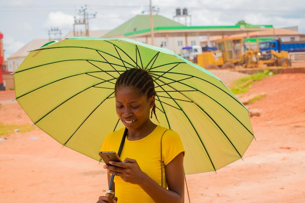 Jovem linda africana usando um guarda-chuva para se proteger sob um clima muito ensolarado e fazendo chamadas com seu celular