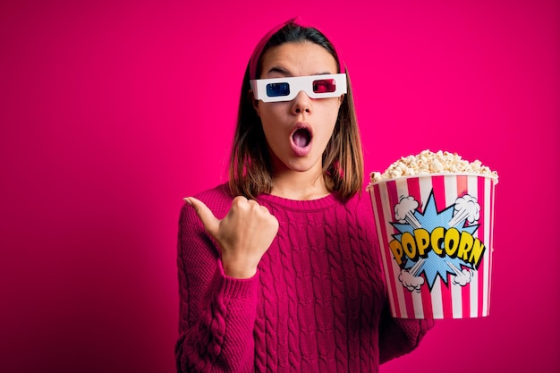 Jovem linda assistindo filme usando óculos 3d comendo caixa com pipocas Surpreso apontando com o dedo da mão para o lado boca aberta expressão espantada