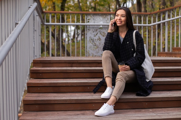 Jovem linda casual com café para falar alegremente no celular nas escadas no parque da cidade