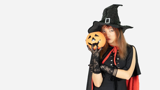 Jovem linda com cabelo preto e laranja em vestido preto e chapéu de bruxa está segurando abóbora no dia de halloweenisolada em fundo branco