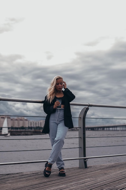 Jovem linda com longos cabelos loiros olhando para o mar e tomando café