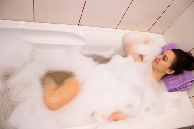 Jovem linda com pacote na cabeça relaxando no banho cheio de espuma no banheiro