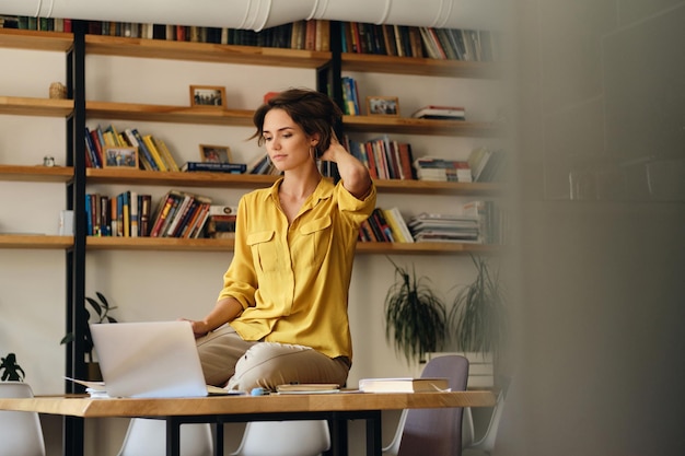 Jovem linda de camisa amarela sentada na mesa enquanto pensativamente trabalhando no laptop no escritório moderno